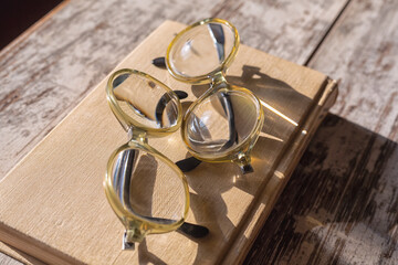 A pair of identical reading glasses lie on top of a closed book in bright evening sunlight on a wooden table, close-up
