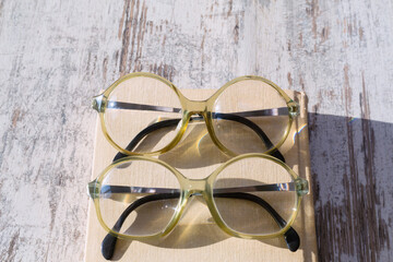 A pair of identical reading glasses lie on a book in bright sunlight on a wooden table, close-up, top view.  literature blogs, advertising bookstores or libraries, articles about vision, eye health