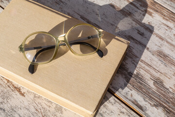 Reading glasses with high optical power lenses lying on a book in bright sunlight on a wooden table, close-up, top view.