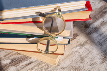 Vintage reading glasses hooked by the temple on the top book and hanging on a stack of books with light reflections on the lenses, wooden background, close-up.