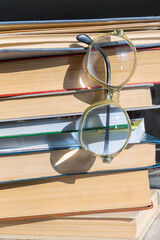 Vintage reading glasses hooked by the temple on the top book and hanging on a stack of books with light reflections on the lenses, close-up, vertical shot.