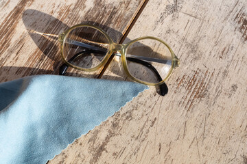 Glasses with strong diopter lenses for reading next to a microfiber lens cleaning cloth on a wooden table surface, close-up.