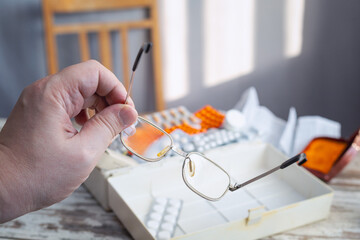 A hand holds glasses against a background of scattered medicines on a table from an open first aid kit, close-up. For illustrating articles about treatment of poor vision, myopia and hyperopia