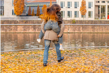 Young happy couple walking playfully canal in modern European city, man giving woman piggyback ride city canal bank, Playful autumn date, partnership and support, Urban romance, modern architecture