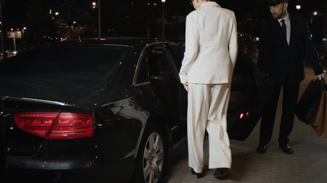 Successful woman getting into back seat of premium class car while male personal driver helping client with opening door and loading shopping bags into vehicle on city street during evening