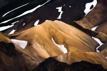 View of rugged, multicolored hills, where golden hues meet dark browns and stark white snow patches, creating a dramatic contrast in Landmannalaugar, Rangarbing ytra, Iceland.