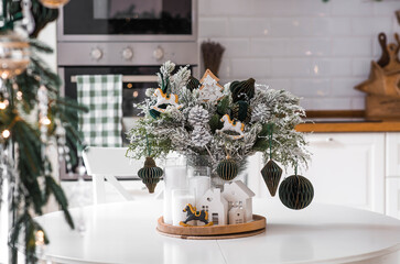 Home decoration for the New Year 2026. A bouquet of fir trees with green toys, horses, and white houses on the kitchen table. Christmas decorations in the interior.