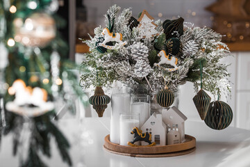 Home decoration for the New Year 2026. A bouquet of fir trees with green toys, horses, and white houses on the kitchen table. Christmas decorations in the interior.