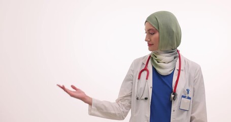 Muslim woman doctor extends open palm presenting space for copy. Medical specialist wears hijab and stethoscope inviting patient to get service - Powered by Adobe