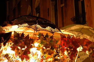 A glowing Venetian mask display at night, showcasing ornate leaf-inspired designs and lace parasols. A vibrant glimpse into Venice’s carnival artistry and festive charm.