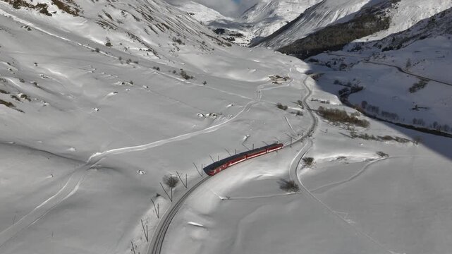 A Red Passenger Train is Running through Snowy Mountain in the Swiss Alps. Aerial View at sunny Day.