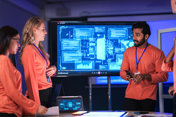 A diverse tech event team in matching orange uniforms collaborates in a high-tech control room, coordinating logistics by analyzing real-time data on a tablet and smartphone.