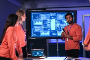 A diverse tech event team in matching orange uniforms collaborates in a high-tech control room, coordinating logistics by analyzing real-time data on a tablet and smartphone.