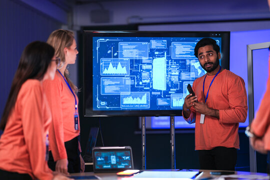 A diverse tech event team in matching orange uniforms collaborates in a high-tech control room, coordinating logistics by analyzing real-time data on a tablet and smartphone.