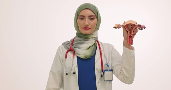 Muslim gynecologist holds anatomical model of uterus and condom on white background. Doctor explains safe methods of contraception. Slow motion