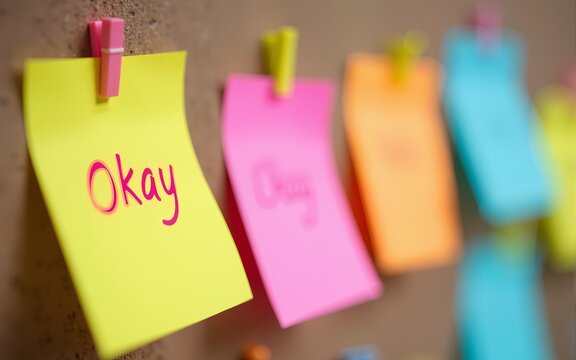 Colorful sticky notes on corkboard. One note says Okay Workplace mental health initiative. Employee support. Caring, encouraging work environment. Positive message for colleagues. Promoting