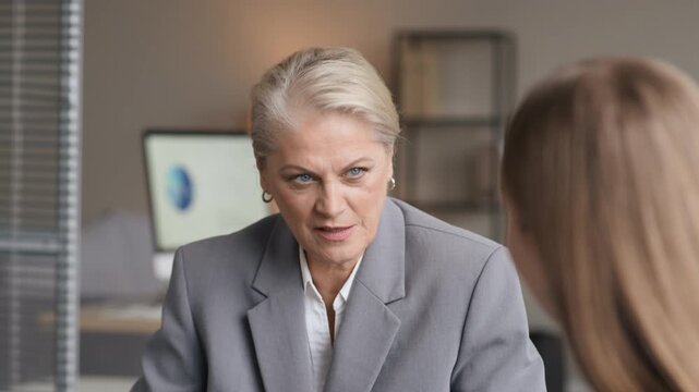 Tilt up shot of confident mature Caucasian female boss sitting at head of conference table and answering questions of female employee during office meeting