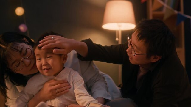 Tender moment of Asian family of three relaxing in warmly lit cozy room in evening, father gently patting his young son head while mother watching lovingly