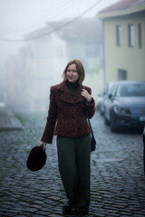 Woman standing on a foggy cobblestone street, holding a hat, dressed in a textured brown jacket and green trousers, with soft morning mist wrapping the quiet neighborhood.