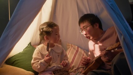 Asian father and his little daughter sitting inside cozy teepee tent playing toy guitar together in warm evening light - Powered by Adobe