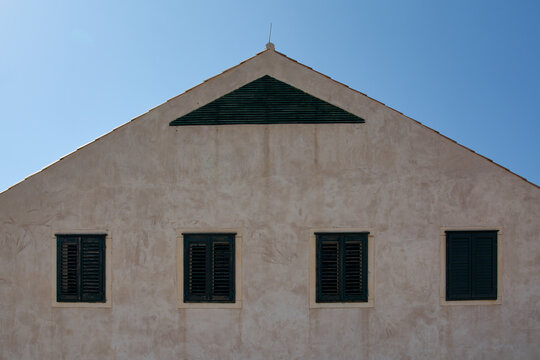 Symmetrical facade of a historic building in Dubrovnik. Four windows with green shutters and a triangular gable against a clear blue sky.