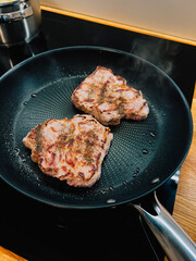 Two seasoned pork neck steaks are frying in hot pan on modern induction stove, creating delicious aroma and steam, classic culinary scene. Sizzling pork neck steaks browning in cast iron skillet