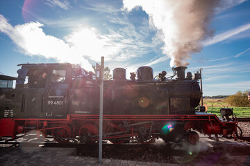 The historic "Rasender Roland" narrow-gauge steam locomotive stands at a rural station on the island of R&uuml;gen, Germany. Steam billows against a clear blue sky, framed by vibrant autumn foliage.