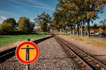 A striking yellow and red warning sign prohibiting access stands prominently on the narrow-gauge railway tracks on R&uuml;gen, framed by autumn trees and a blue sky.