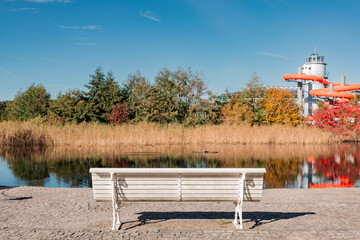 A white bench overlooks a pond reflecting autumn trees and the tower and orange water slides of a modern spa or leisure facility in the background.
