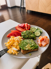 Grey plate with wholemeal avocado toast, scrambled eggs, smoked salmon, sliced tomatoes, strawberries and cucumbers served for breakfast on white table. Wholemeal avocado toast with scrambled eggs