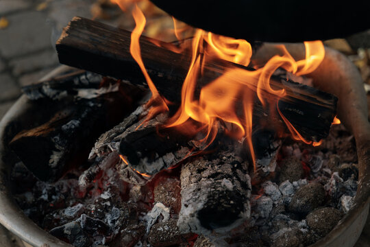 A close-up (macro) of an open fire: a bright orange flame and smoldering red embers burning in a special metal tray. Concept: the element of fire, warmth, combustion, outdoor cooking.