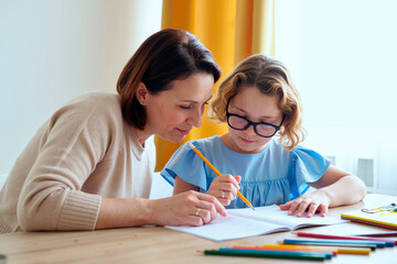 Caucasian middle aged woman helping Caucasian child girl with homework at desk, both focusing on writing in notebook, colored pencils scattered on table, learning together