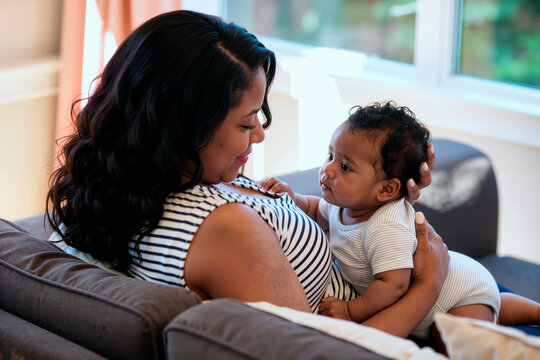 Black woman holding infant child while sitting on sofa, mother gazing lovingly at baby, baby looking up at mother, natural light coming through window in background