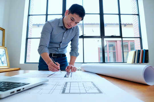 Asian young adult man standing at desk working on architectural blueprints with pencil and ruler, large window in background, laptop and rolled plans on table