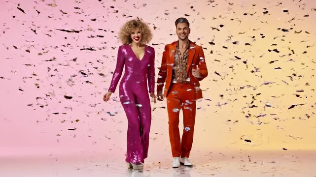 Glamorous couple wearing funky 70s disco outfits celebrating on a colorful background. Energetic man and woman dancing happily as golden confetti falls around them during a festive party