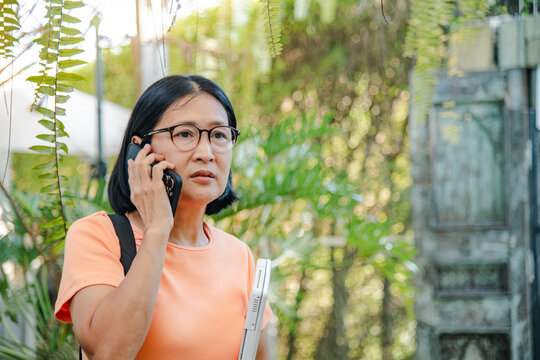 An Asian businesswoman talking on her phone while holding a laptop, seeking a seat in an outdoor garden, a vintage greenery fern cafe, with a refreshing bokeh background, a digital nomad lifestyle