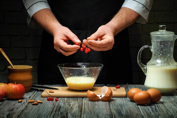 Hands are seen preparing ingredients by cracking eggs and adding berries into a bowl. Nearby are apples, milk, and almonds, creating a warm kitchen atmosphere