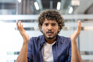 Close-up photo of a worried young Indian man sitting at a desk in an office facing the camera and talking emotionally