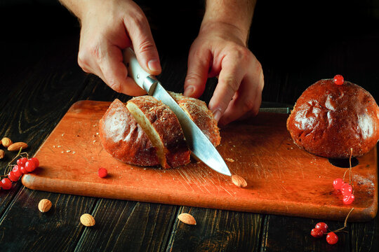Fresh bread is being sliced with a sharp knife on a wooden cutting board. Red berries and almonds are placed around the bread, creating a cozy kitchen atmosphere - Powered by Adobe