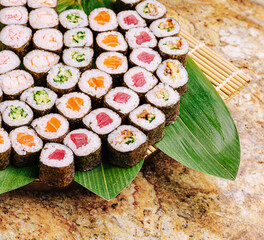 A variety of colorful sushi rolls sit on a bamboo platter, showcasing different fillings and toppings. The platter rests on a stone surface, inviting a culinary experience.