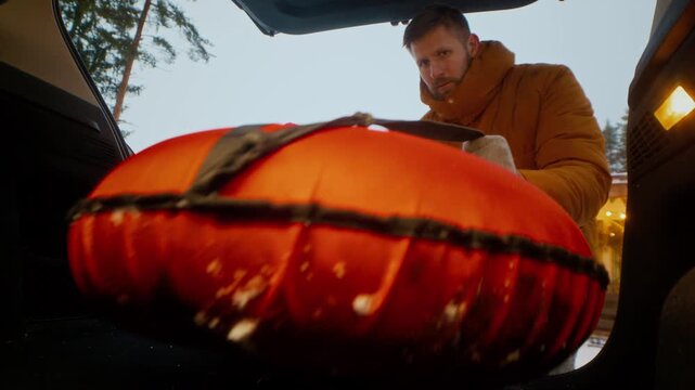 Modern Caucasian family with child placing inflatable sled and ice skates into open car boot preparing for winter holiday trip in quiet countryside