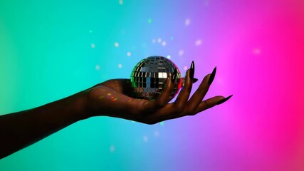 Dark skinned woman's hand with long, manicured nails holding a sparkling disco ball against a vibrant, multi colored background, perfect for a celebration, party, or retro themed event