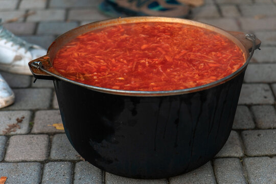 A close-up of a large black metal cauldron (kazan) filled with boiling, bright red thick soup (borscht or goulash). The cauldron sits directly on the gray paving stones. A traditional hot dish.