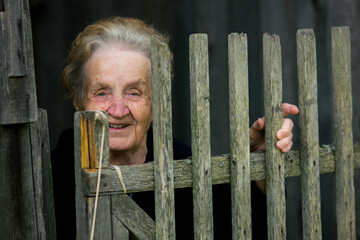 Smiling elderly woman peeks from behind an old wooden fence, her hand gently holding one of the weathered slats.