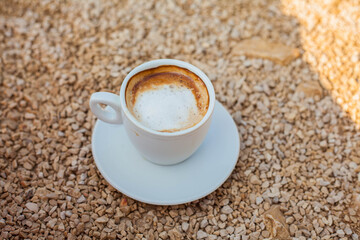 Morning coffee with cream in a white cup on a rocky beach. Warm sunlight, peaceful seaside mood, and a cozy start to the day with natural coastal textures.