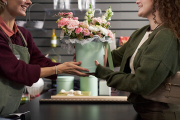 Caucasian young adult woman handing pastel green box, filled with assorted fresh flowers to smiling Caucasian young adult female florist behind counter in flower shop interior