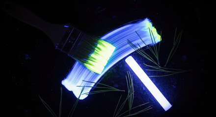 Paintbrush with glowing paint on bristles over dark surface. Pine needles and UV light wand arranged nearby. Phosphorescent paint brush stroke, glowing in dark party theme
