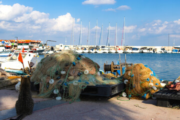 Close-up of a Large Pile of Tangled Commercial Fishing Nets, Ropes, and Buoys on a Dock, with Moored Sailboats and Blue Water in the Background.