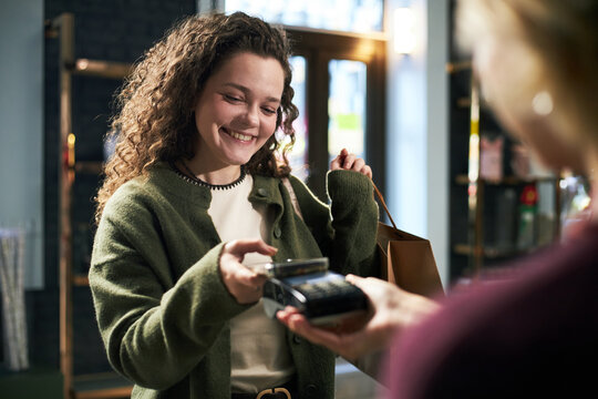Young adult Caucasian woman smiling while making contactless payment with smartphone at checkout counter, holding shopping bag in modern retail store, interacting with cashier