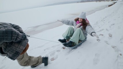 Red cheek boy pulling his young mother on sled along snowy path beside winter forest and lake creating cheerful family moment - Powered by Adobe